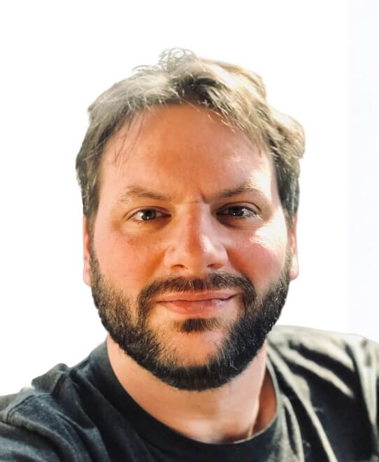 A man with short brown hair and a beard looks at the camera with a slight smile. He is wearing a dark t-shirt, set against a bright background—perfectly reflecting the approachable vibe of Team V2 tech leads.