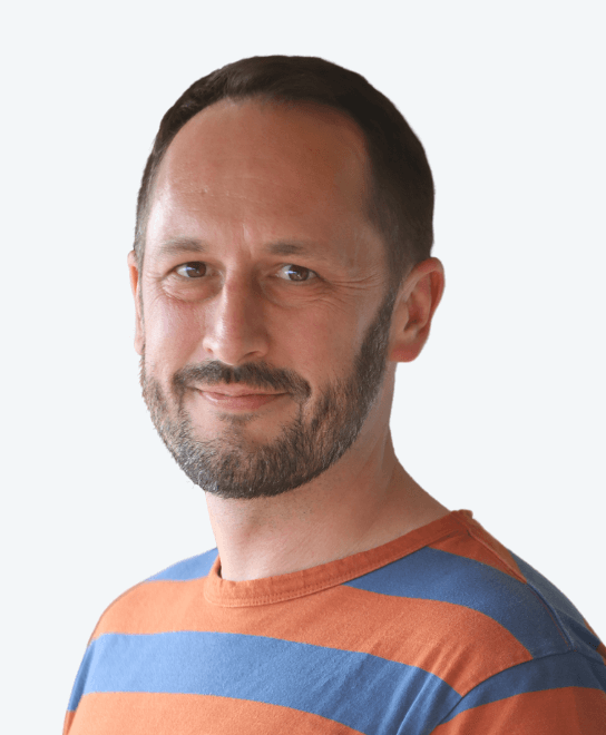 A man with short brown hair and a beard, wearing an orange and blue striped shirt, smiles slightly while facing the camera against a plain light background, embodying Team V2’s spirit of leadership.