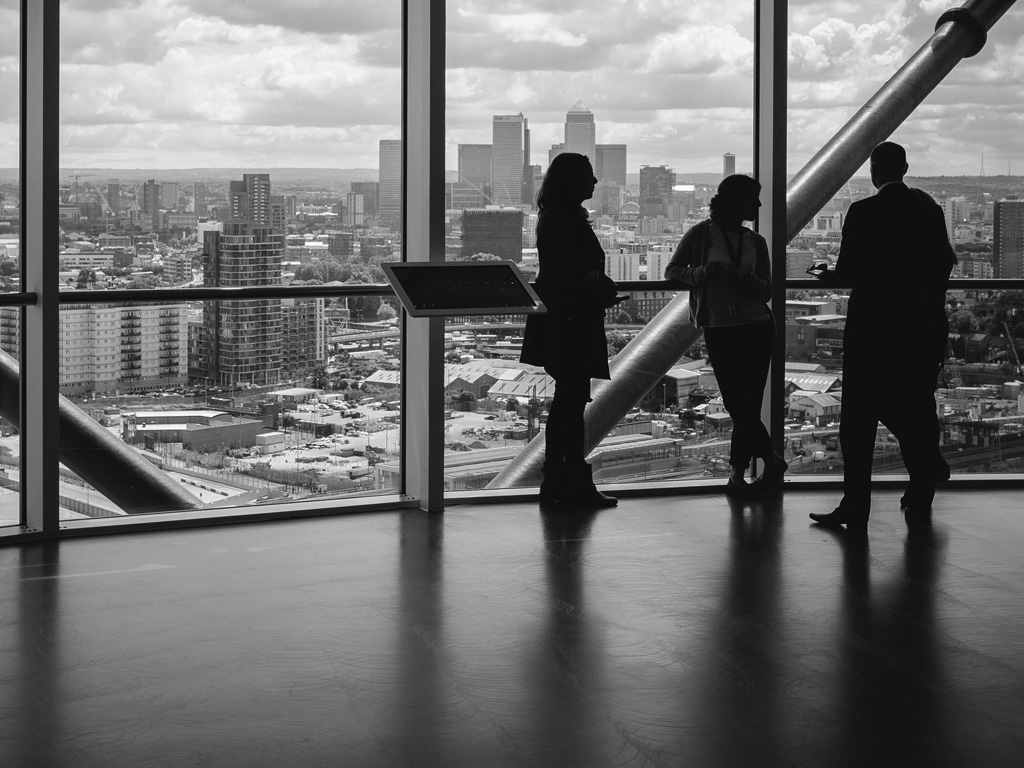 Three business-people looking out skyscraper at city buildings in a black and white photo. Photo by Charles Forerunner on Unsplash.