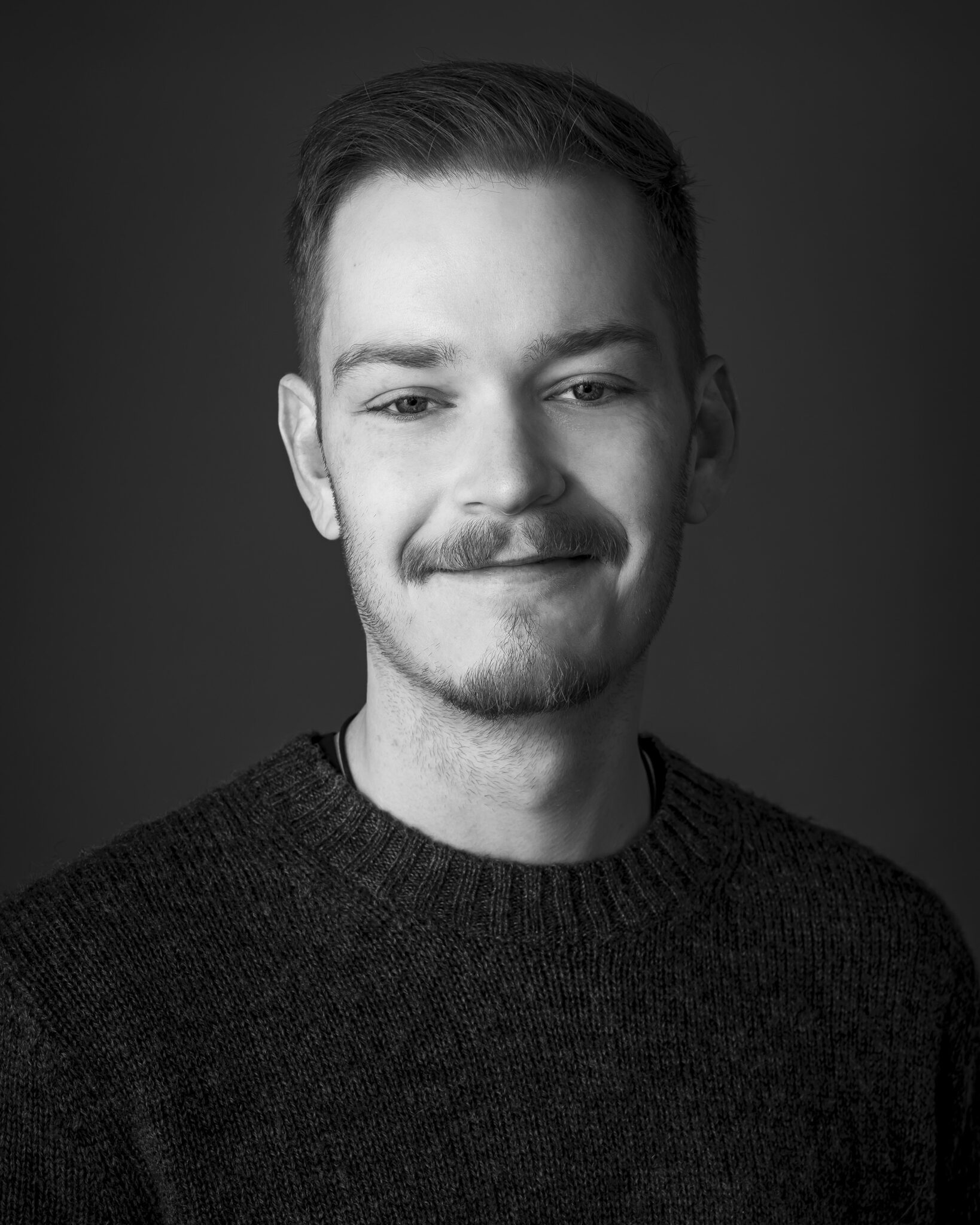 A young man with short hair and a trimmed beard, wearing a textured sweater, poses for a portrait against a plain dark background.