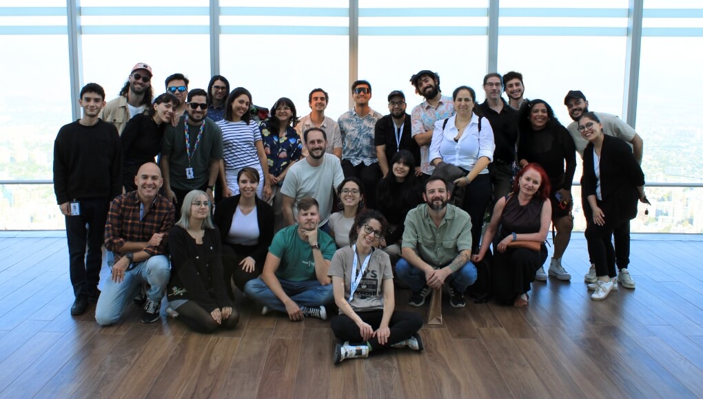 A group of around 28 people pose together inside a bright room with large windows and a city view, all smiling at the camera.