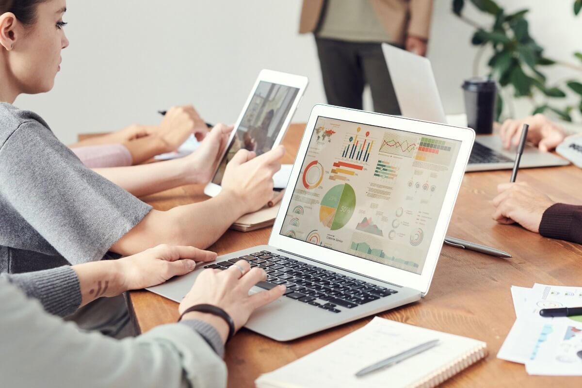 People sit at a table with laptops, tablets, and papers, analyzing colorful charts and graphs while sharing success stories during a business meeting.