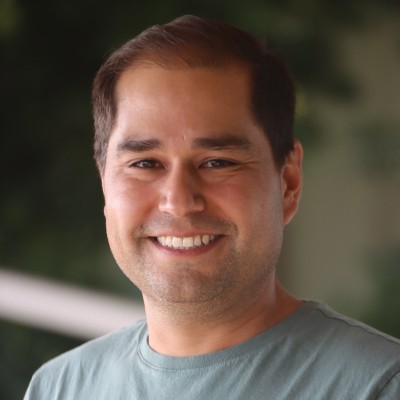 A man with short dark hair smiles at the camera, wearing a green t-shirt, with a blurred background of greenery, featured in the 2025 Annual Report.