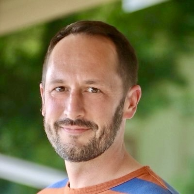 A man with short brown hair and a beard is smiling slightly, wearing a blue and orange shirt, with the 2025 Annual Report reflected in the blurred green background.