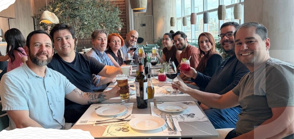 A group of ten people sit around a restaurant table, smiling and holding drinks, celebrating their Year in One Idea with empty plates and bottles on the table in front of them.