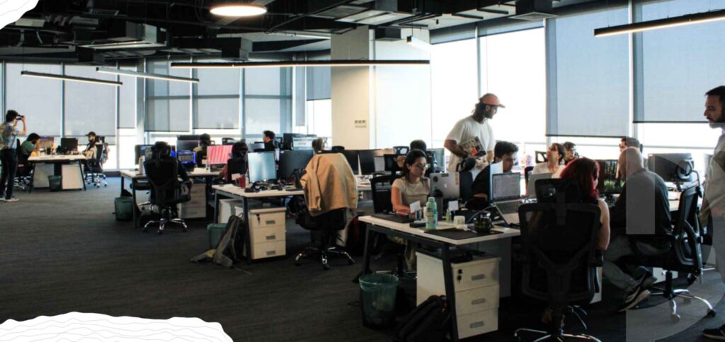 Large open-plan office with several people working at desks, computers, and monitors under ceiling lights, next to large windows with blinds.