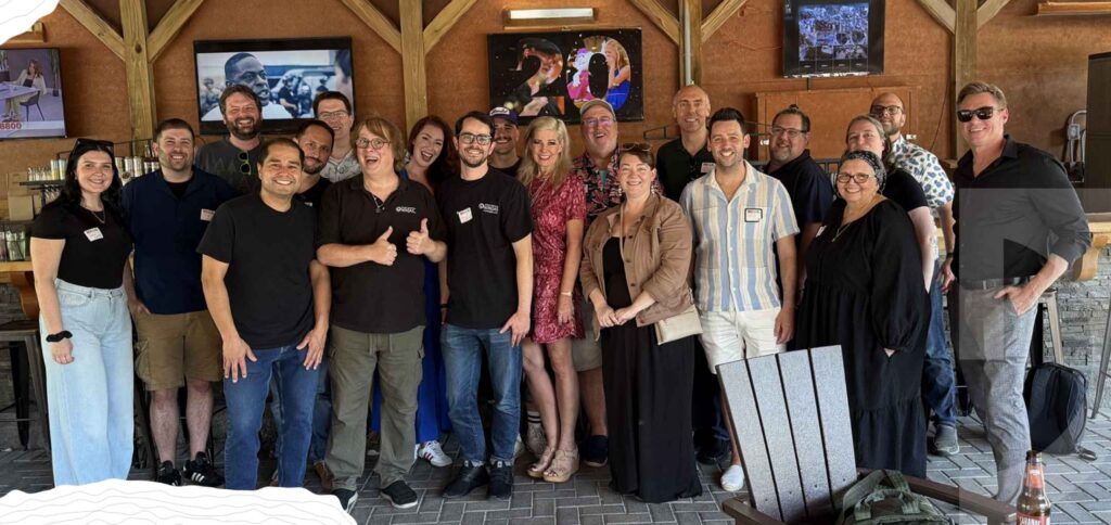 A group of people posing and smiling together at an outdoor bar or lounge, with multiple TVs and wooden beams in the background, celebrating the release of the 2025 Annual Report.