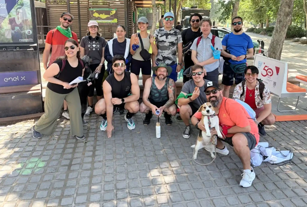 A group of people pose outdoors on a cobblestone path, some standing and others kneeling, with one person holding a small dog. The gathering marks the launch of the 2025 Annual Report, with trees and park structures visible in the background.