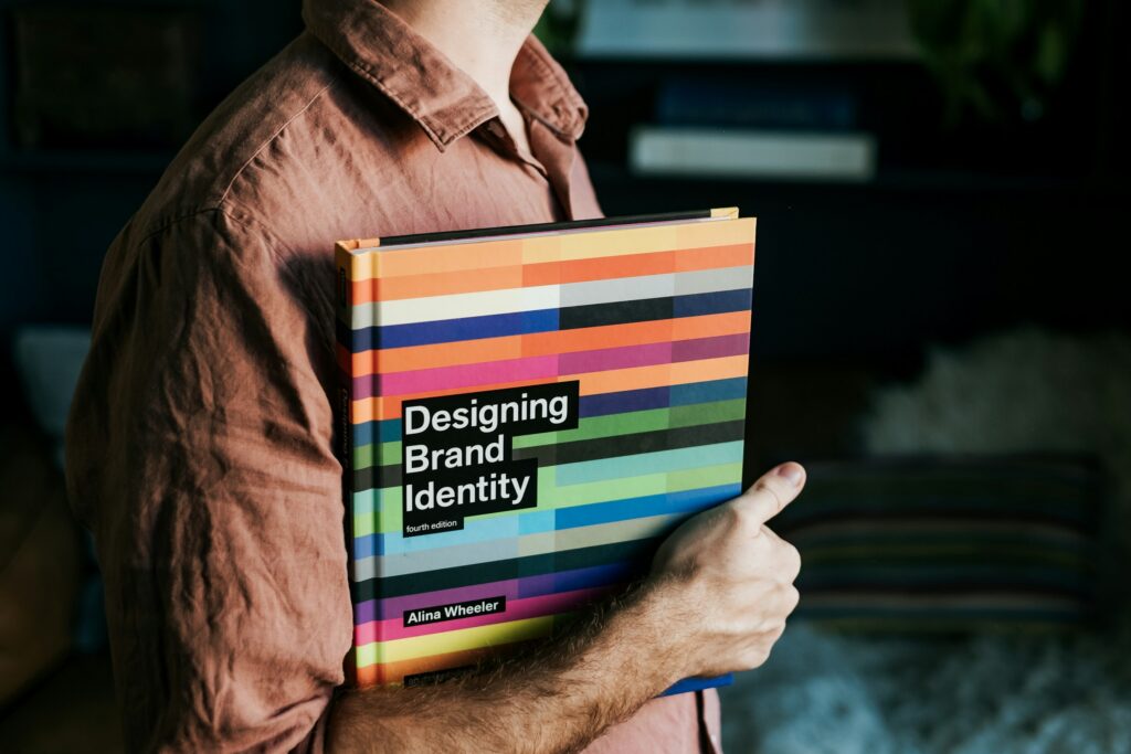 A person in a brown shirt holds a copy of "Designing Brand Identity" by Alina Wheeler, featuring a colorful striped cover, while filling out a Brand Source Map worksheet.