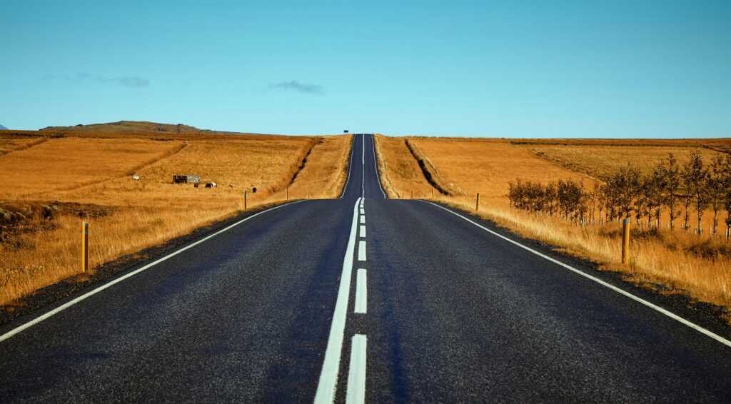 A straight, empty asphalt road with white lines stretches into the distance through dry, golden fields under a clear blue sky, symbolizing the journey of long-term SEO and its compounding effect for sustained organic ROI.