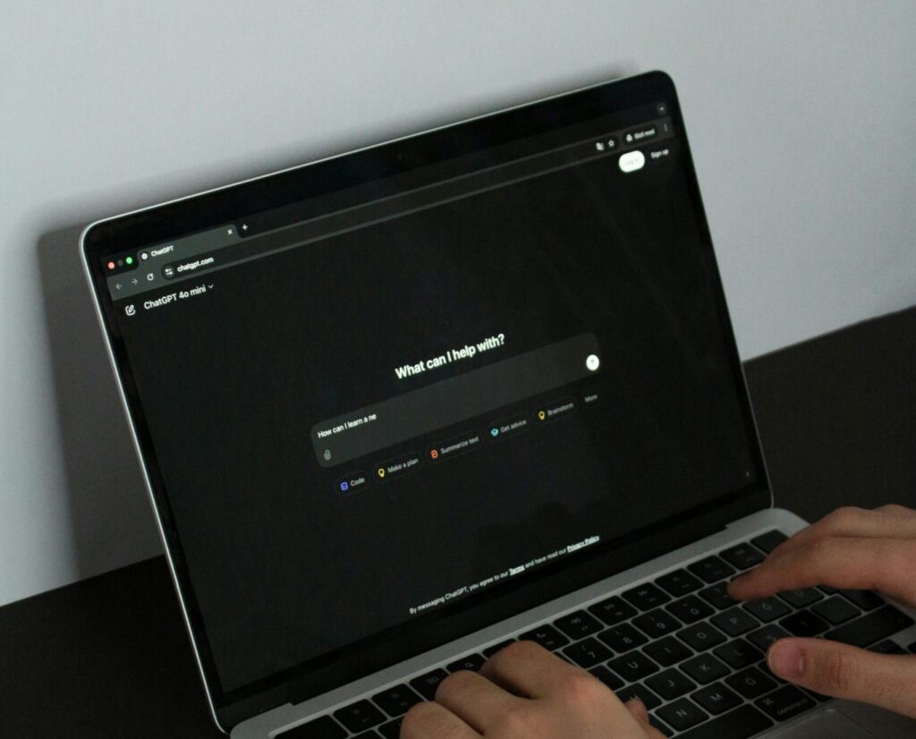 Person typing on a laptop with a dark screen displaying a search bar that says, "What can I help with?"—perfect for boosting organic visibility or managing product feeds on a sleek black desk.