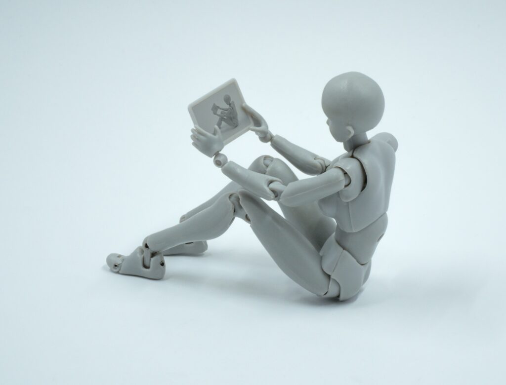 A gray humanoid figurine is sitting on the ground, holding and looking at a small rectangular tablet—perhaps exploring AI search optimization on its screen.