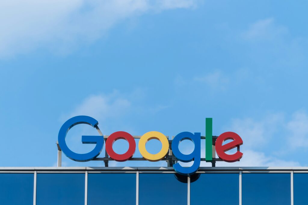 The Google logo in colorful letters is displayed on top of a glass office building against a blue sky background, representing Google's commitment to AI Search Optimization and innovation.