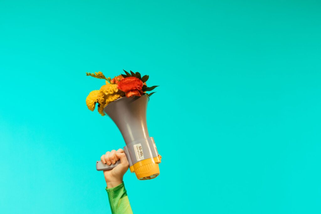 A hand holds a megaphone filled with colorful flowers against a bright turquoise background.